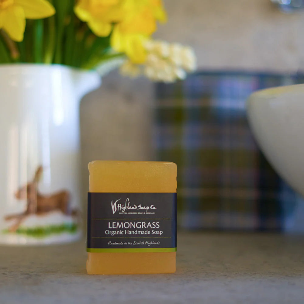 Handmade soap bar with 'Highland Soap Company' branding in front of a blurred background with flowers and a mug